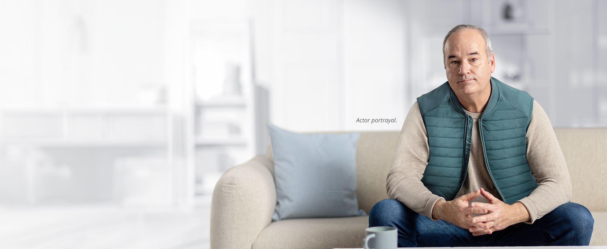 An older man sitting on a beige couch with his hands clasped together, looking forward with a serious expression. The footnote next to him reads: Actor portrayal.