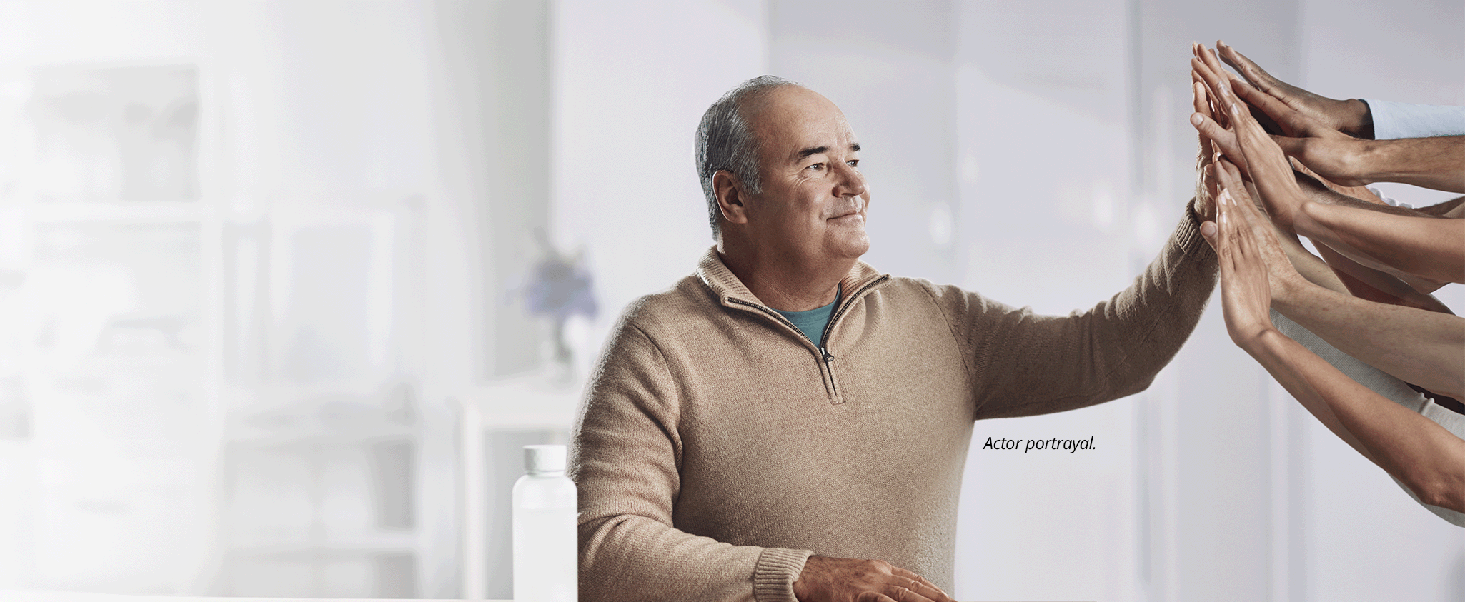 An older man stands in a white room, smiling into the distance. He has his hand raised in a high-five, and many hands reach out from the side to return the high-five. The footnote next to him reads: Actor portrayal.