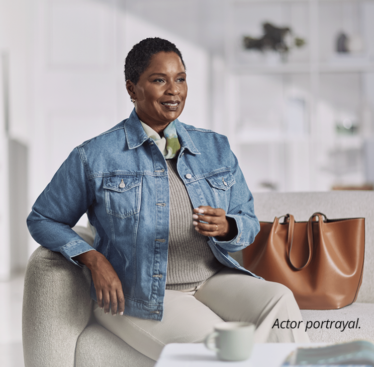 An older woman sits on a white couch, wearing a beige outfit and jeans jacket with a brown leather tote next to her. She's relaxed, gently smiling into the distance. Disclaimer reads Actor portrayal.